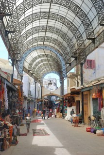 A vibrant market street with ornate metal canopy, bustling with people and colorful merchandise.