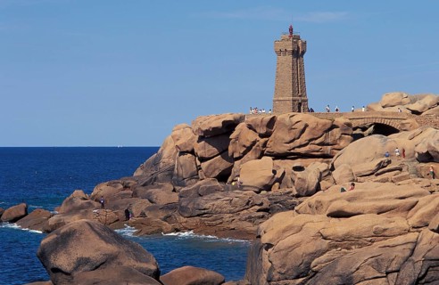 A lighthouse stands on a rocky coastline with people nearby and a clear blue sky. The granit rose