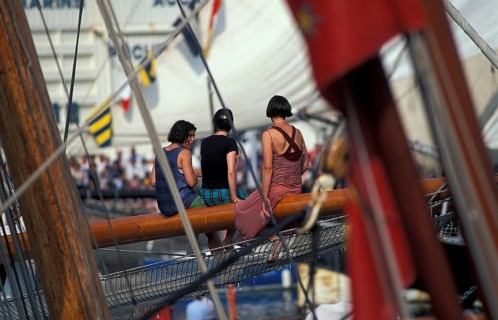 Three women sitting on a sailboat's wooden deck surrounded by nautical flags and ropes. Maritime