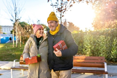 Elderly couple holding christmas presents smiling and walking in a city park during a sunny winter