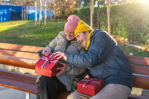 Elderly couple exchanging christmas gifts while sitting on a bench in a city park during a sunny
