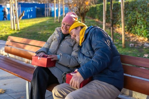 Smiling senior couple exchanging christmas gifts while sitting on a bench in a winter city park