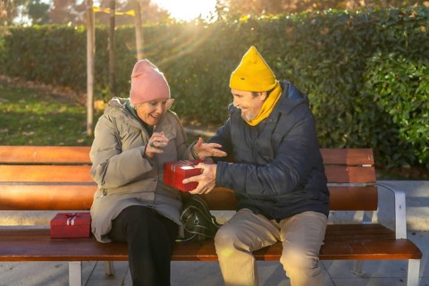 Elderly man surprising his wife with a festive christmas gift while sitting together on a park