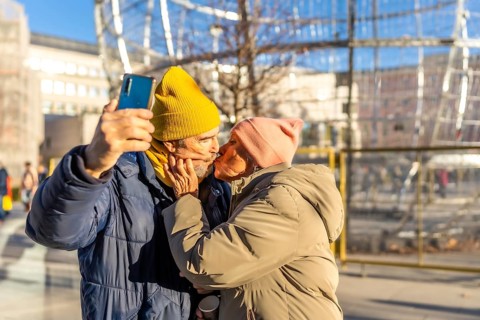Happy senior couple kissing and taking a selfie with smartphone in a winter city during christmas