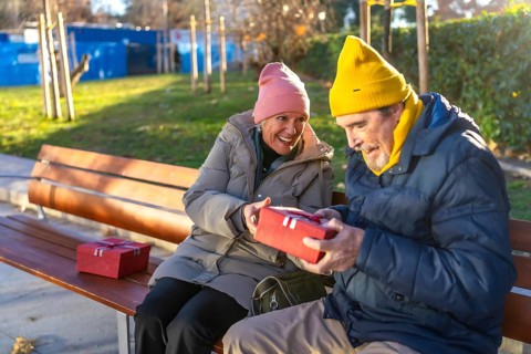 Elderly man receiving christmas gift from smiling wife while sitting on bench in city park