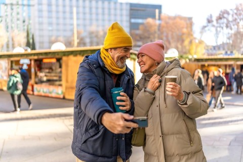 Smiling senior couple taking a selfie and drinking hot beverages at a christmas market in the city