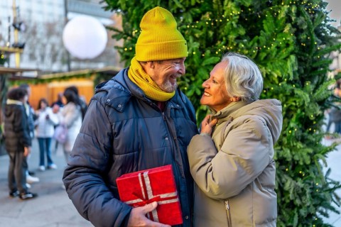 Elderly man surprising his wife with a christmas present at a christmas market during winter