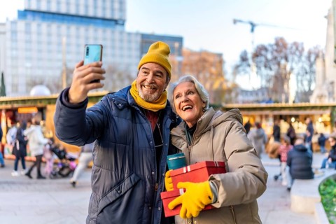 Cheerful senior couple taking a selfie with smartphone at a christmas market, holding gifts and