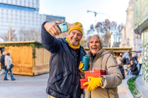 Elderly couple taking a selfie with smartphone at a christmas market, holding gifts and hot drinks,