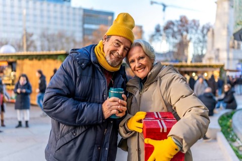 Smiling senior couple holding christmas gift and hot beverage at outdoor christmas market, enjoying