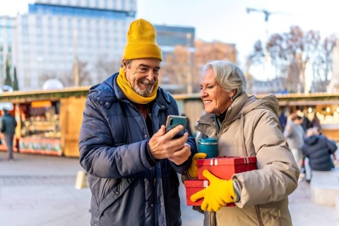 Happy retired couple looking at smartphone and holding christmas gifts and reusable coffee cup at a