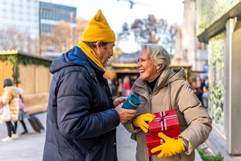 Smiling elderly couple exchanging christmas presents and enjoying hot beverages at a winter market,