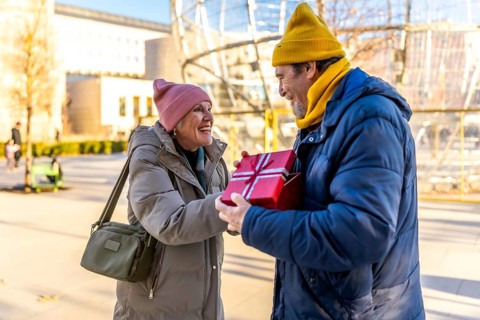 Elderly man joyfully giving a christmas present to a smiling woman in a sunny winter city park,