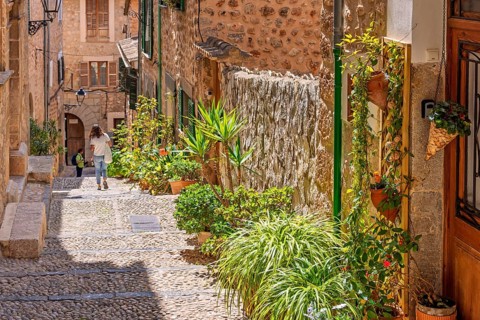 Alone woman walking in an idyllic cobblestone alley in an old village with green plants and flowers