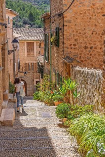 Woman walking in an idyllic cobblestone alley in an old village with green plants and flowers in