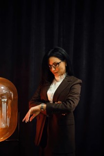 A woman dressed in a fashionable brown suit stands near a glowing lamp. She checks her watch with a