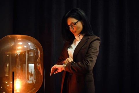 A confident woman in a tailored brown suit stands beside a stylish lamp. She checks her watch