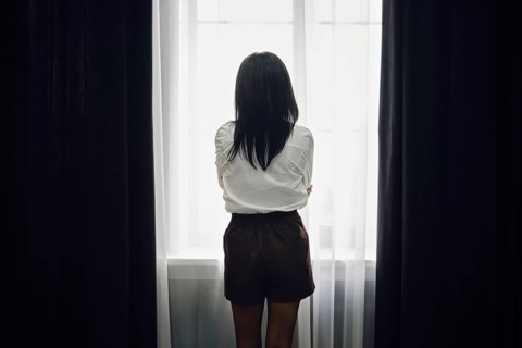 A woman stands with her back to the viewer, gazing out of a bright window. The soft afternoon light