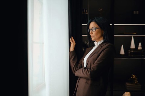 A professional woman dressed in a brown suit stands thoughtfully by a window, gazing outside