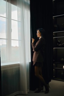 A woman dressed in a tailored suit stands by a large window, pulling back sheer curtains. She