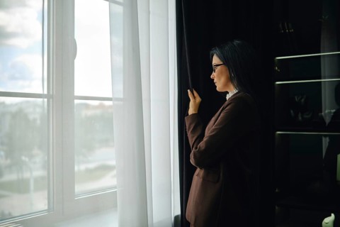 Woman in brown jacket gazes out window while reflecting on the day in a modern setting