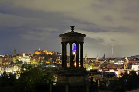 Night scene of Edinburgh with illuminated buildings and a historical monument in the foreground,