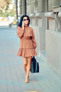 A fashionable woman strolls down the city sidewalk wearing a brown dress and sunglasses, carrying a