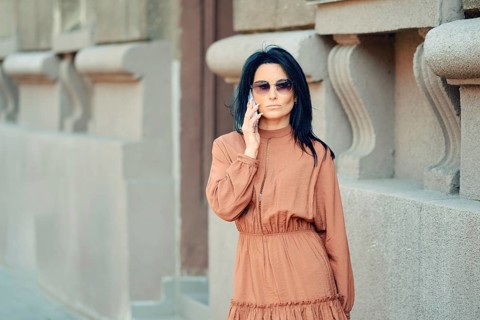 A woman wearing a brown dress and sunglasses engages in a phone conversation while passing by the