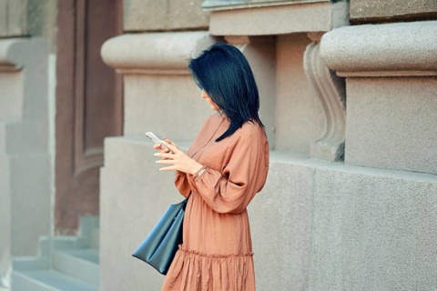 A woman in a flowing brown dress and a handbag stands against a stylish urban building. She is