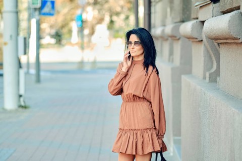 A fashionable woman dressed in a brown dress engages in a phone conversation while standing on a