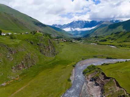 River flowing through green valley, surrounded by mountains and clouds, near a small village,