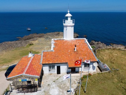 A lighthouse with terracotta roof on a rocky coast with flags and a wide ocean in the background,