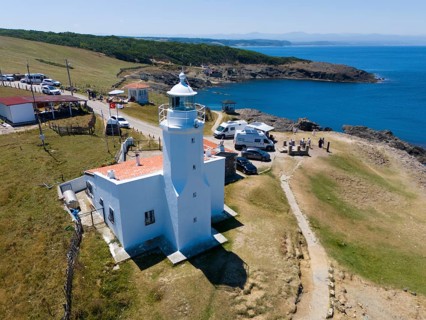 A lighthouse on the coast with mobile homes nearby, surrounded by meadow and rocks, the sea horizon