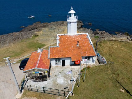 A lighthouse on a rocky coast with blue sky and sea, next to it a building with a terracotta roof,
