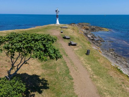 Path leading to a lighthouse on the coast, surrounded by trees and benches in a summer landscape,