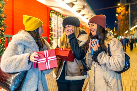 Three cheerful women exchanging christmas gifts and enjoying hot beverages at a vibrant night
