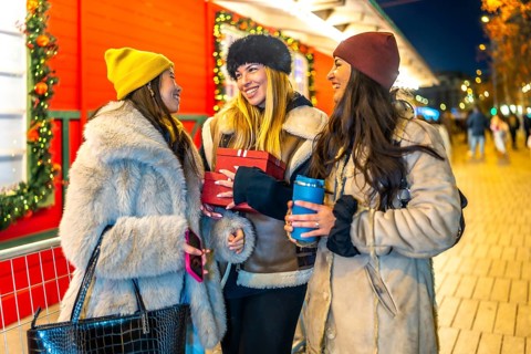Three cheerful women holding christmas gifts and hot drinks, enjoying christmas shopping at a