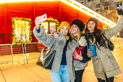 Three happy women taking selfie with smartphone in front of illuminated christmas market kiosk