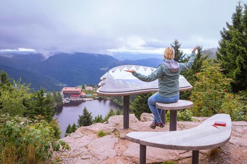 Woman sitting on a wooden bench at a viewpoint, overlooking a lake and mountains, Mummelsee,
