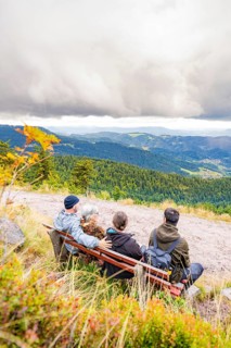 Three people on a bench enjoy the wide view over wooded mountains and valleys, Mummelsee, Seebach,