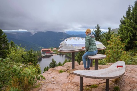 Woman at the panoramic viewpoint with information board, view of lake and forest under clouds,