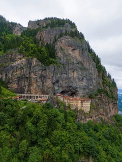 A monastery perched monumentally on a steep rock face, surrounded by dense forests, aerial view,