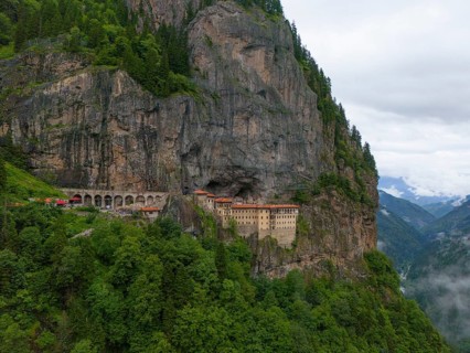 A monastery embedded in a steep rock face surrounded by lush forests and mountains under an