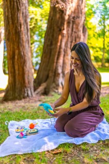 Happy young latin mother kneeling on a blanket in a park, preparing vibrant baby toys while