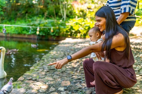 Mother and baby daughter pointing at birds by the pond in a botanical garden, enjoying a sunny day