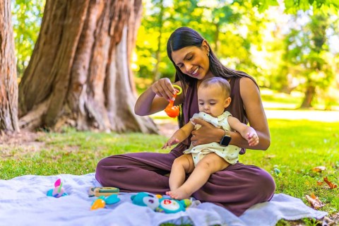 Latin mother holding her baby daughter and playing with colorful toys while sitting on a blanket in