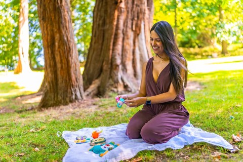 Smiling latin mother arranging baby toys on a blanket in a park, enjoying a sunny day outdoors