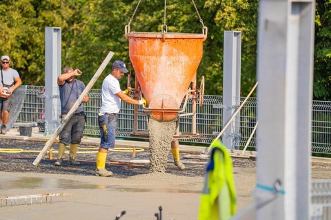 Two workers pouring concrete from large mixer tubs, surrounded by safety measures and mesh fencing,
