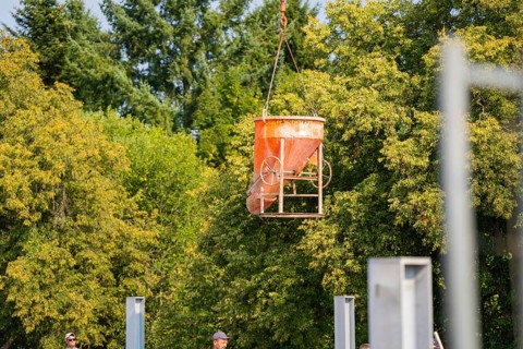 An orange-coloured concrete bucket hovers over a building site. Trees and a clear sky in the