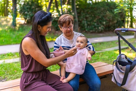 Mother feeding her baby girl solid food, with grandmother holding her, sitting on a park bench
